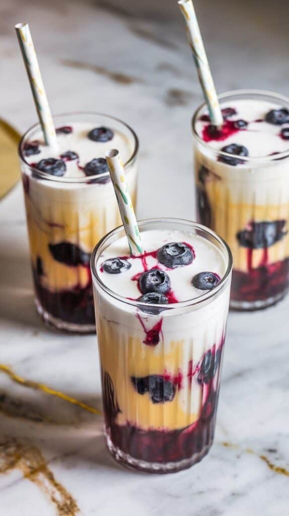 two blueberry muffin dirty sodas served in clear tall glasses with visible layers of cream soda, blueberry syrup, and sweet cream, each with a paper straw