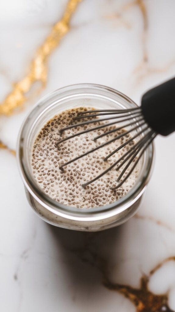 coconut milk, chia seeds, maple syrup, and vanilla extract being whisked together in a small clear jar, texture starting to thicken with chia seeds evenly dispersed