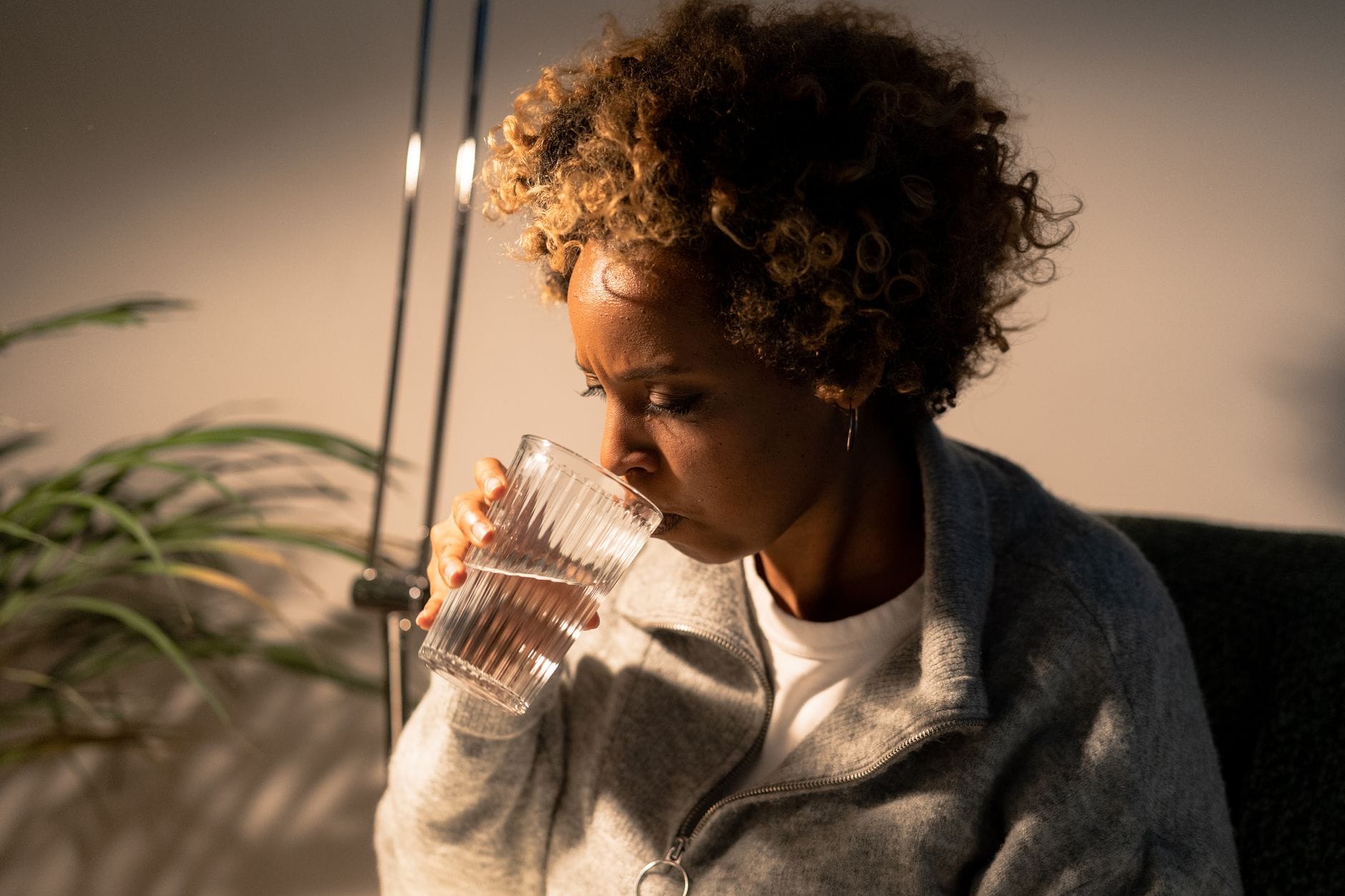 Young woman in a white top sipping water from a clear glass near a bright window