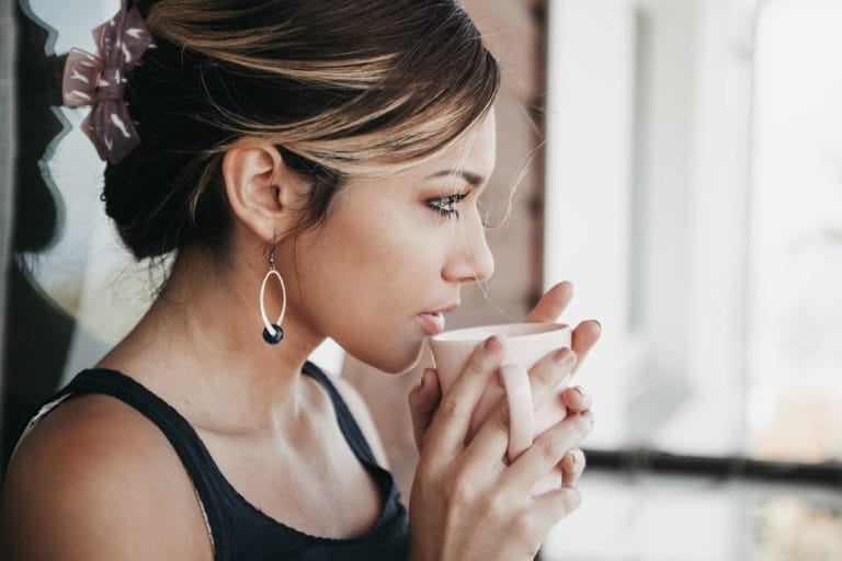 Young woman wrapped in a blanket holding a steaming mug of tea by a window