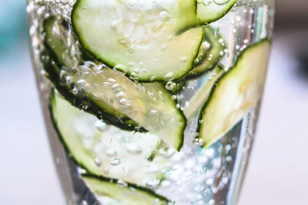 Tall glass of water with cucumber slices, mint, and ice on a bright counter