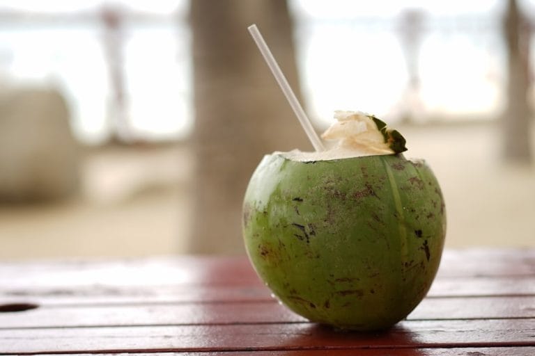 A glass of pale coconut water with a striped straw beside a halved green coconut
