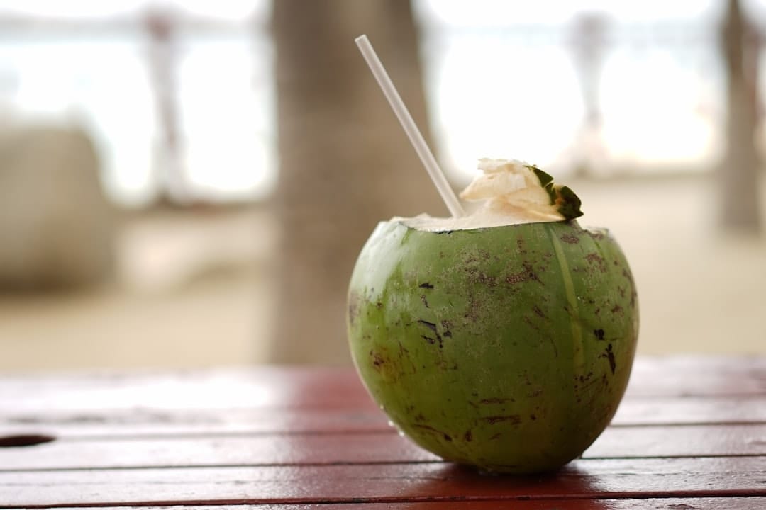 A glass of pale coconut water with a striped straw beside a halved green coconut